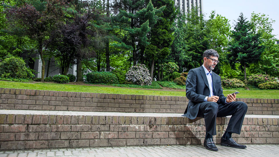 A man in a suit sits on a ledge in a park looking at his phone