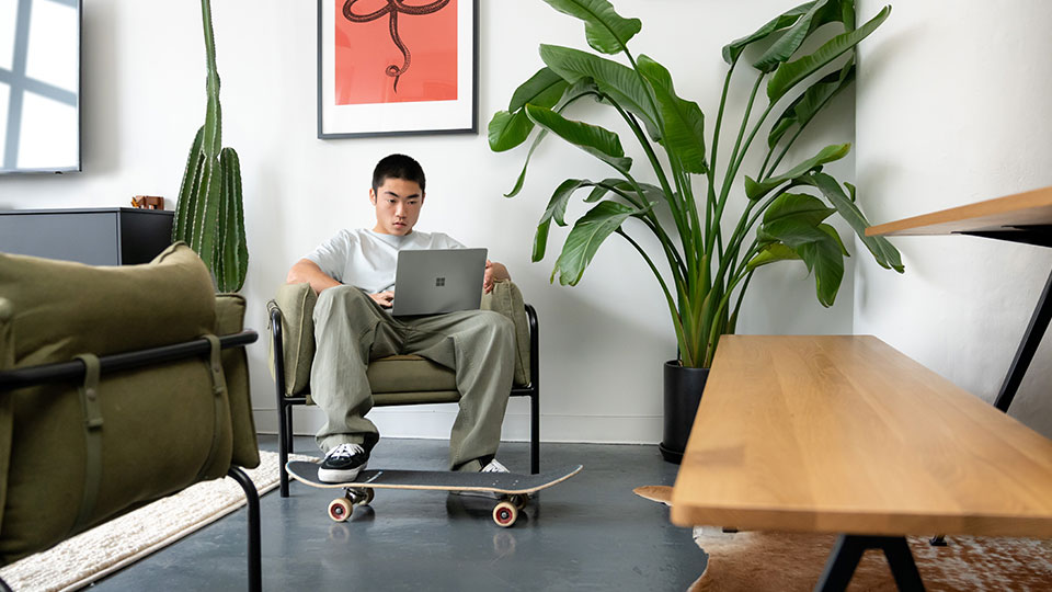 A man sits in a chair with one foot balanced on a skateboard while he works on a laptop on his lap